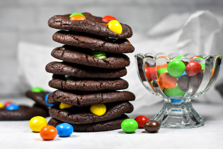 A stack of chocolate M&M cookies and a glass bowl of M&Ms