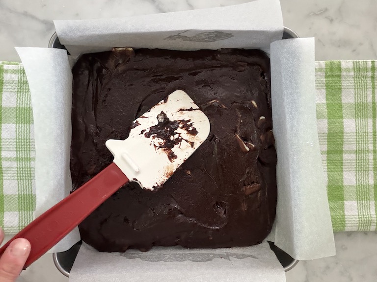 spatula spreading brownie batter into a parchment lined tin