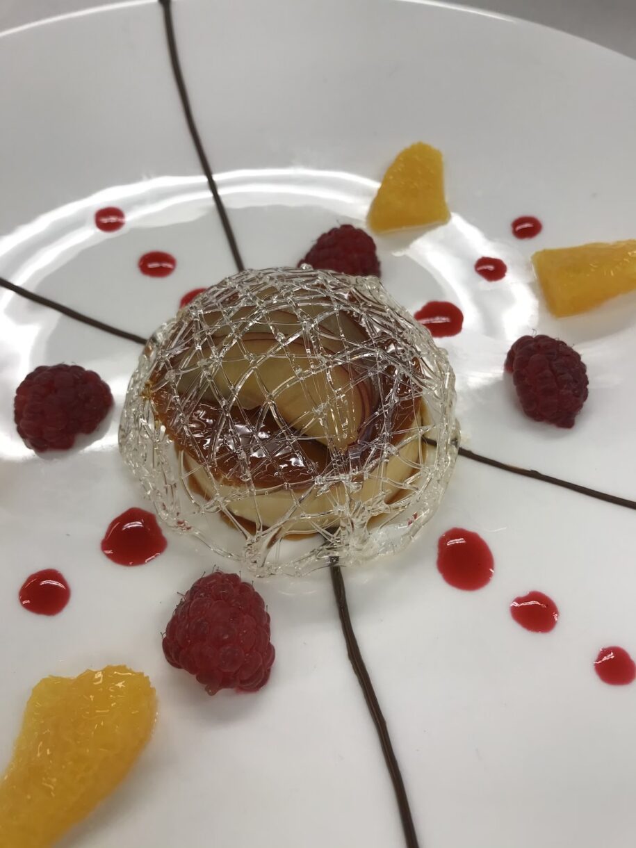 Creme caramel with sugar cage, fruit coulis, and chocolate piping, on a white plate
