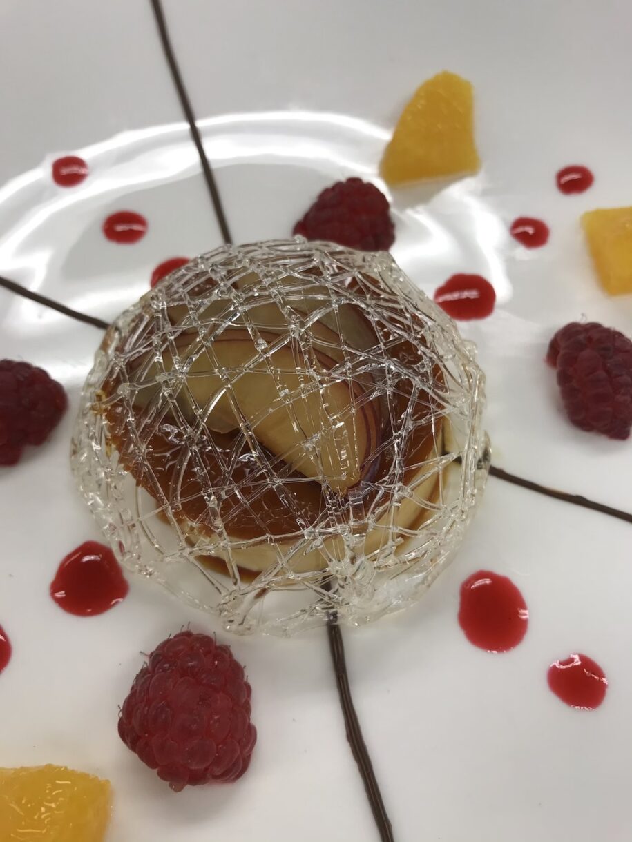 A closeup of the sugar cage surrounding creme caramel, plated on white plate with fruit coulis piping