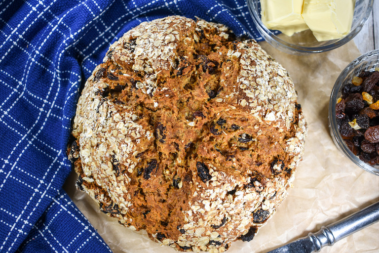 A loaf of soda bread with a blue tea towel and dishes of butter and mixed dried fruit