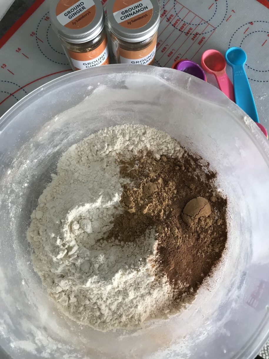 A mixing bowl with flour and spices, jars of ginger and cinnamon, and measuring spoons