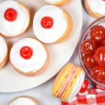 A white plate with bakewell macarons, a bowl of cherries, and gingham towel