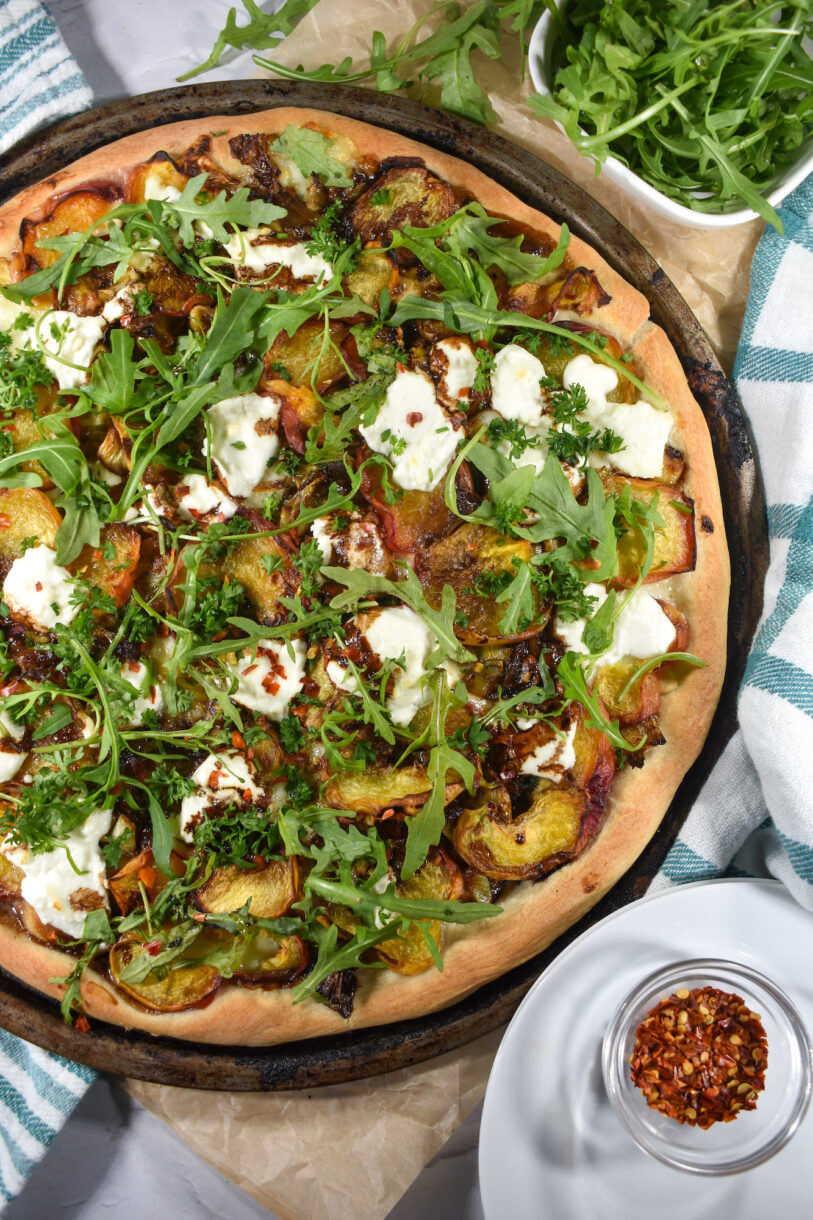 A pizza with peaches, goat cheese, mozzarella, and arugula, and small bowl of chili flakes