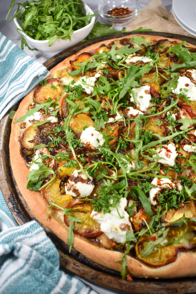 Peach and goat cheese pizza on white surface with tea towel, plates, and bowl of pepper flakes and arugula