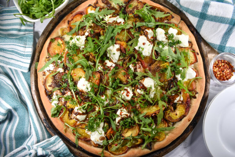 Peach and goat cheese pizza on white surface with tea towel, plates, and bowl of pepper flakes and arugula