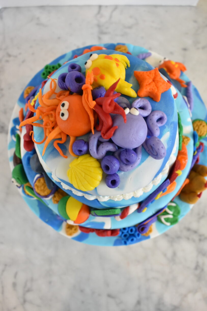Octopus, fish, and seashell decorations on top of a beach themed fondant cake