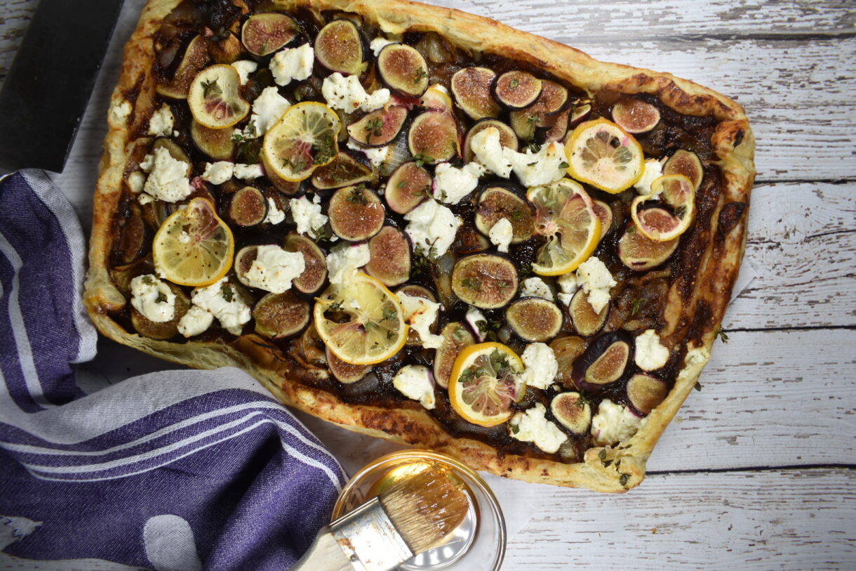 Puff pastry fig galette on a white wood background, along with a bowl of honey and a purple tea towel