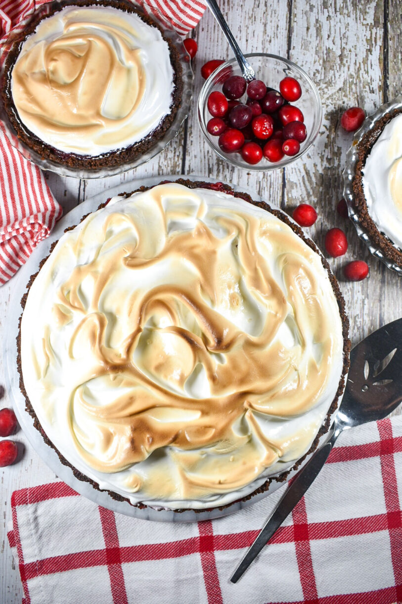 Meringue pie on a white wood surface with a bowl of cranberries, and a red and white tea towel