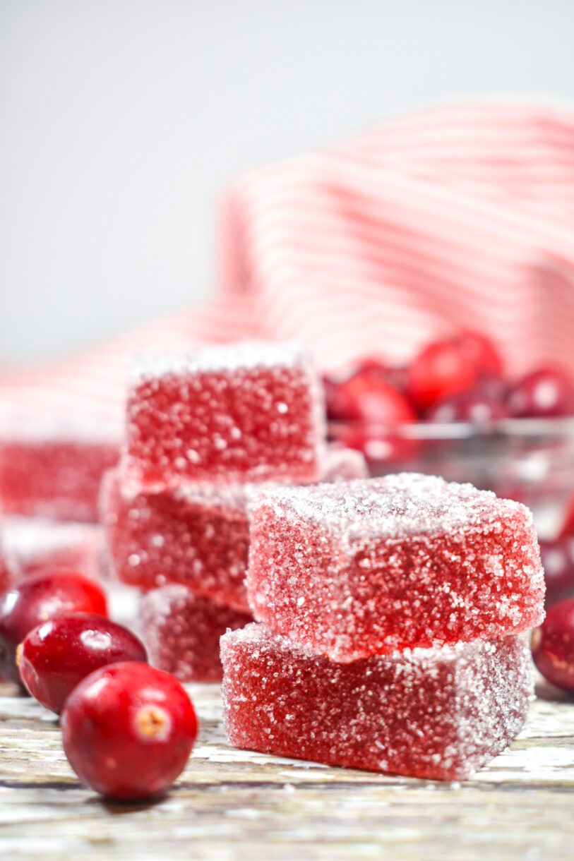 Squares of cranberry jelly candy and fresh cranberries on a white surface, with red tea towel behind