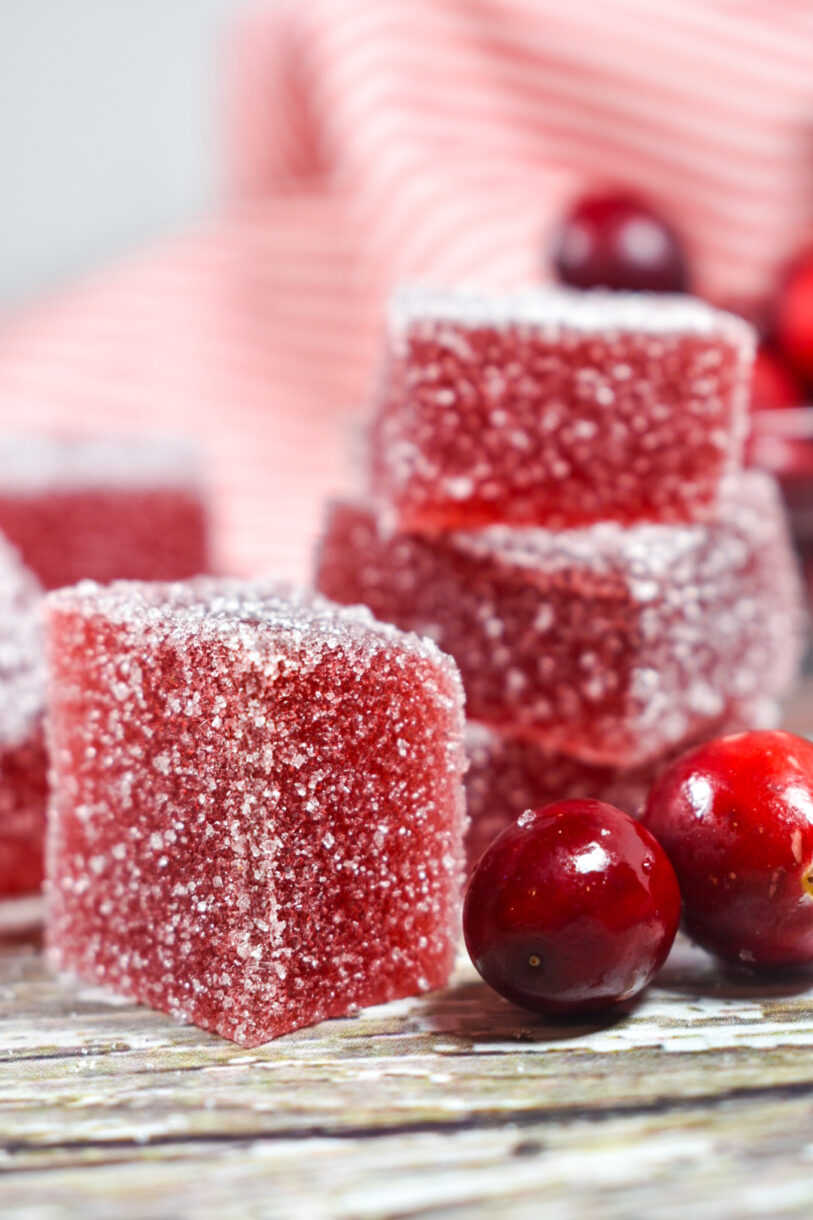 Squares of cranberry jelly candy and fresh cranberries on a white surface, with red tea towel behind