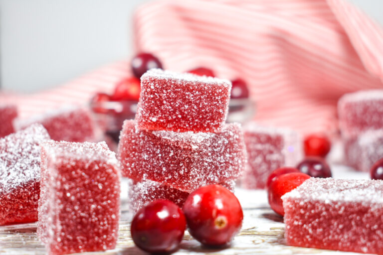 Squares of cranberry jelly candy and fresh cranberries on a white surface, with red tea towel behind