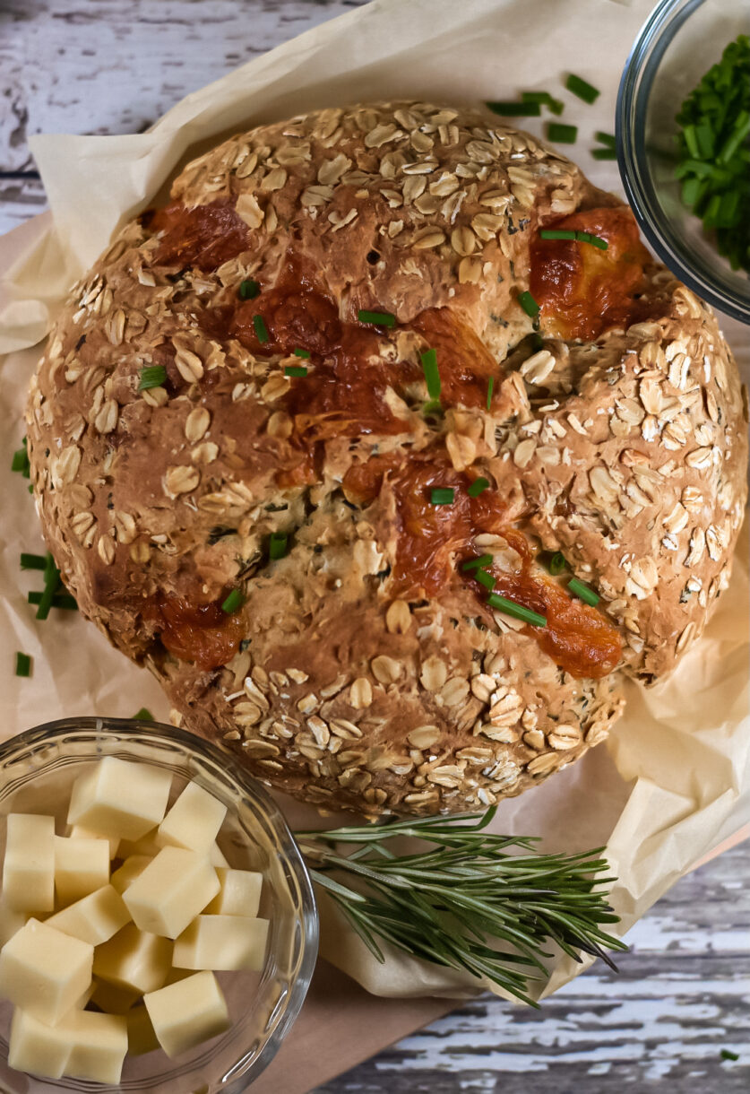 A loaf of soda bread on a sheet of parchment with a bowl of cheese cubes and spring of rosemary