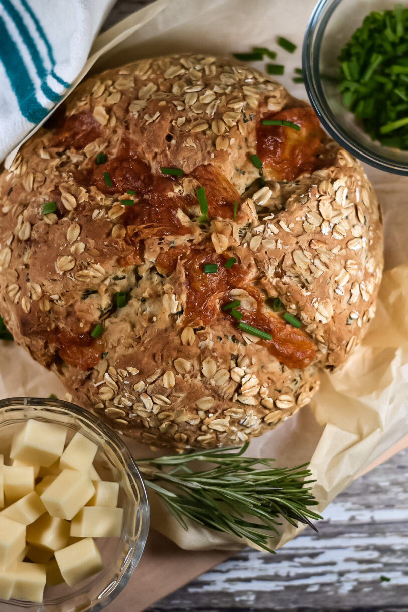 A loaf of soda bread on a sheet of parchment, surrounded by herbs, a bowl of cheese, and a tea towel