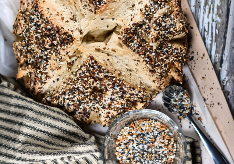 Everything bagel bread, a brown striped tea towel, a dish of seasoning, and a metal spoon