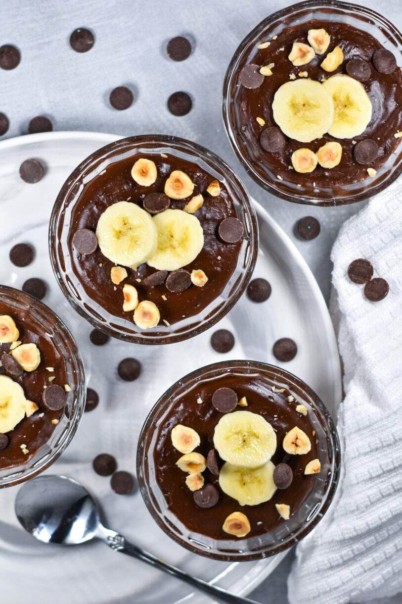 Glass bowls of chocolate pudding garnished with peanuts, bananas, and chocolate chips, sitting on a white plate