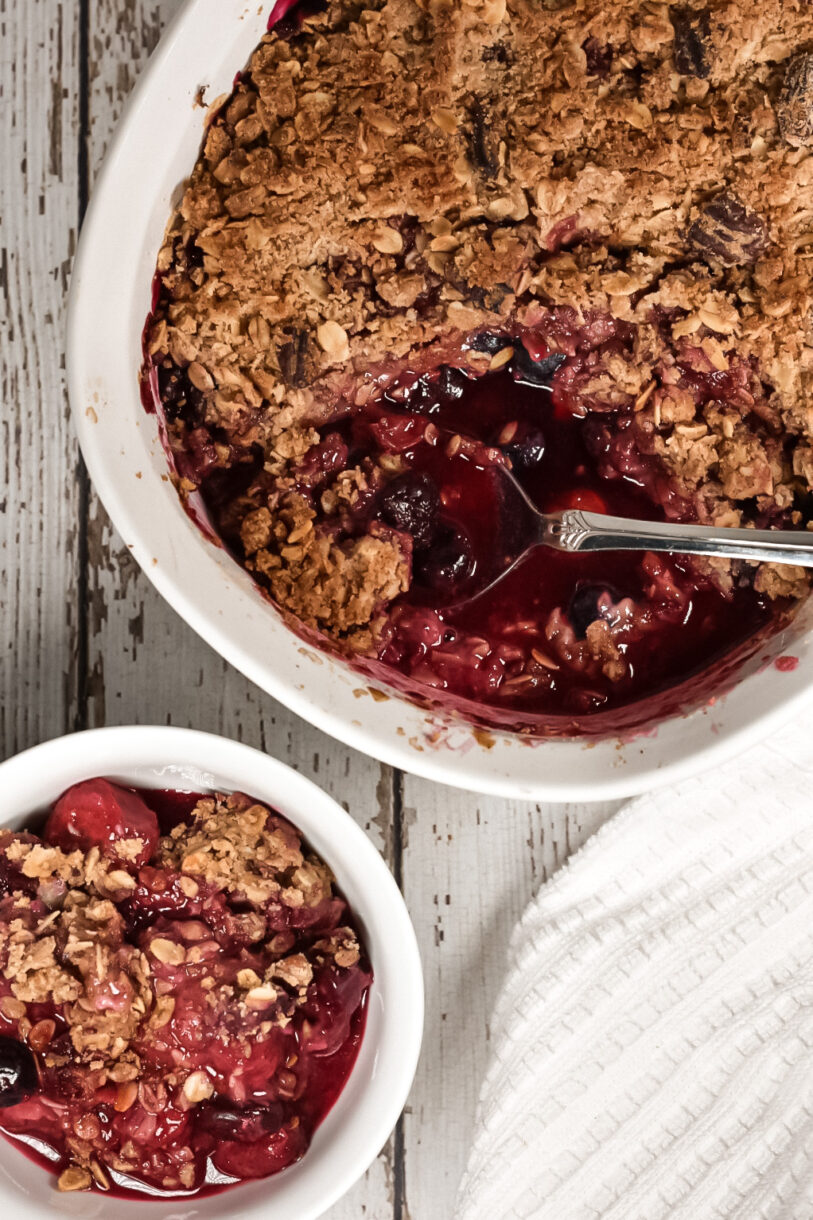 A large casserole dish with a vegan berry crumble and a spoon, plus a portion in a small white dish, on a white wooden background