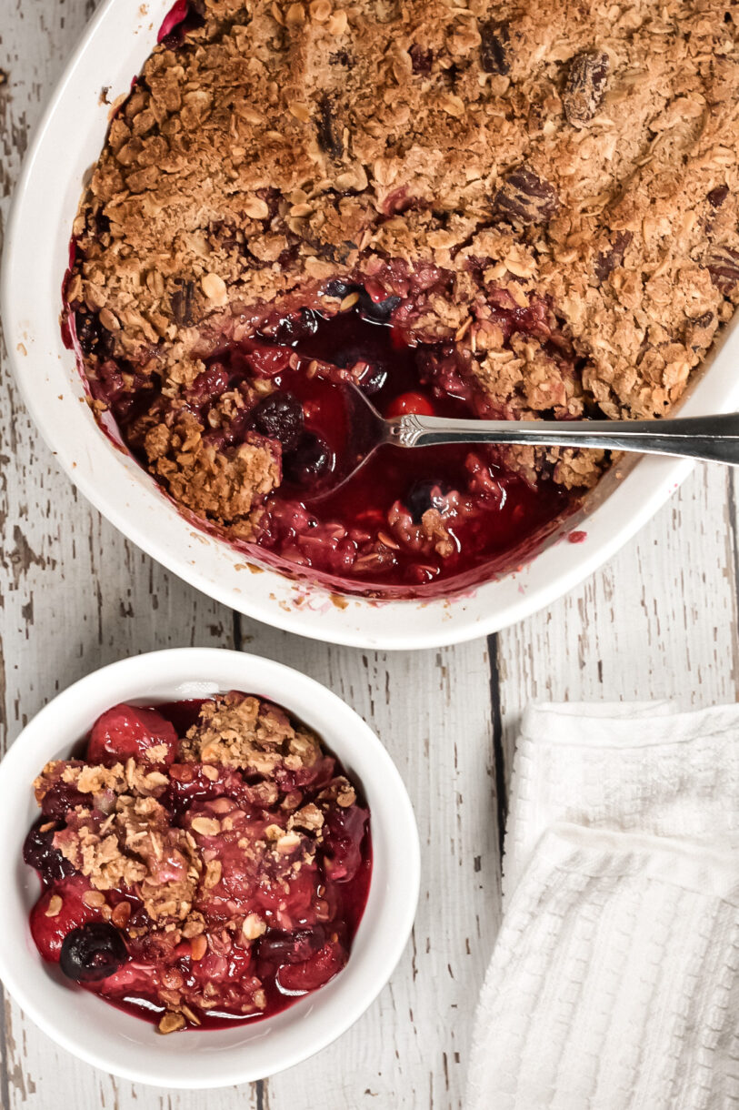 A casserole dish and small dish of vegan berry crumble, with a spoon and white tea towel