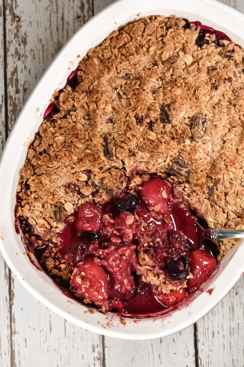 Wooden background and a white oval casserole dish with a vegan berry crumble and a spoon