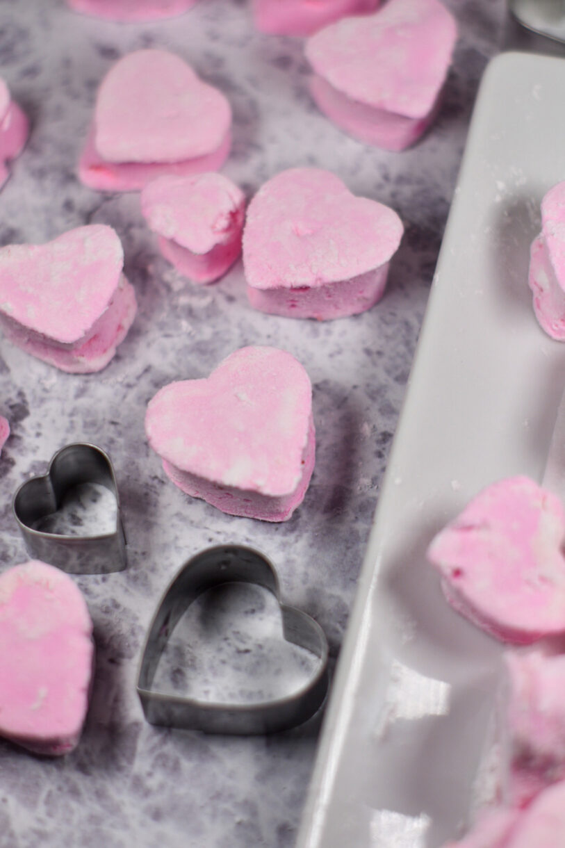 Pink heart shaped strawberry marshmallows and a pair of heart cookie cutters on a grey surface
