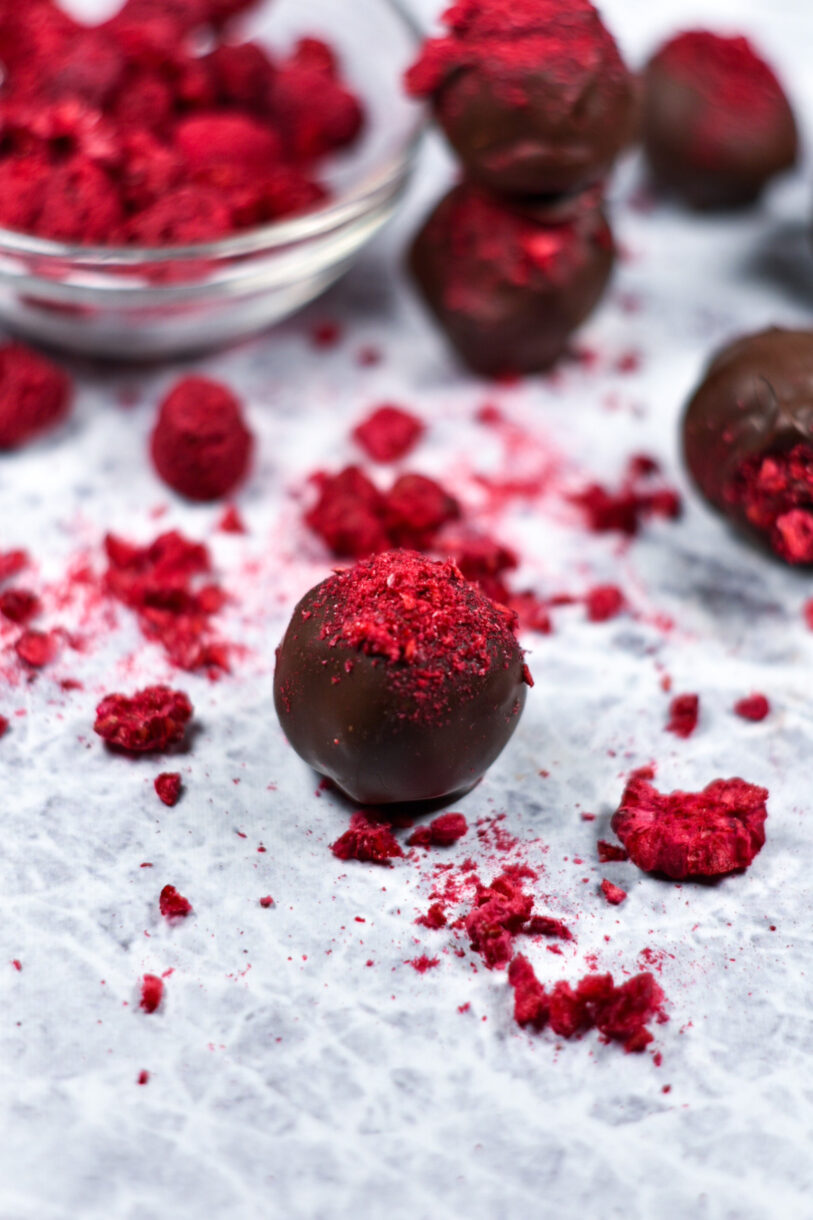 A chocolate truffle garnished with bright red freeze dried raspberries, and a bowl of raspberries in the background