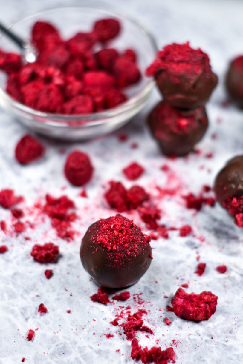 A bowl of vibrant red freeze-dried raspberries and a chocolate raspberry truffle on a white surface