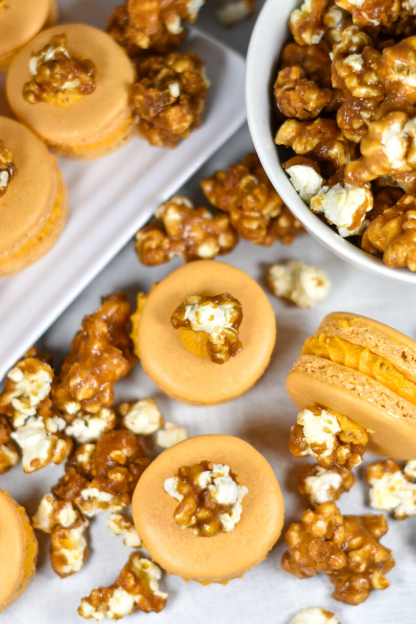 Caramel popcorn buttercream macarons surrounded by popcorn, on a white surface