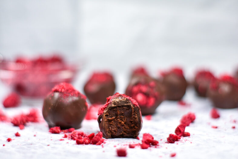 Horizontal shot of a chocolate ganache truffle with a bite missing, surrounded by crushed dried raspberries