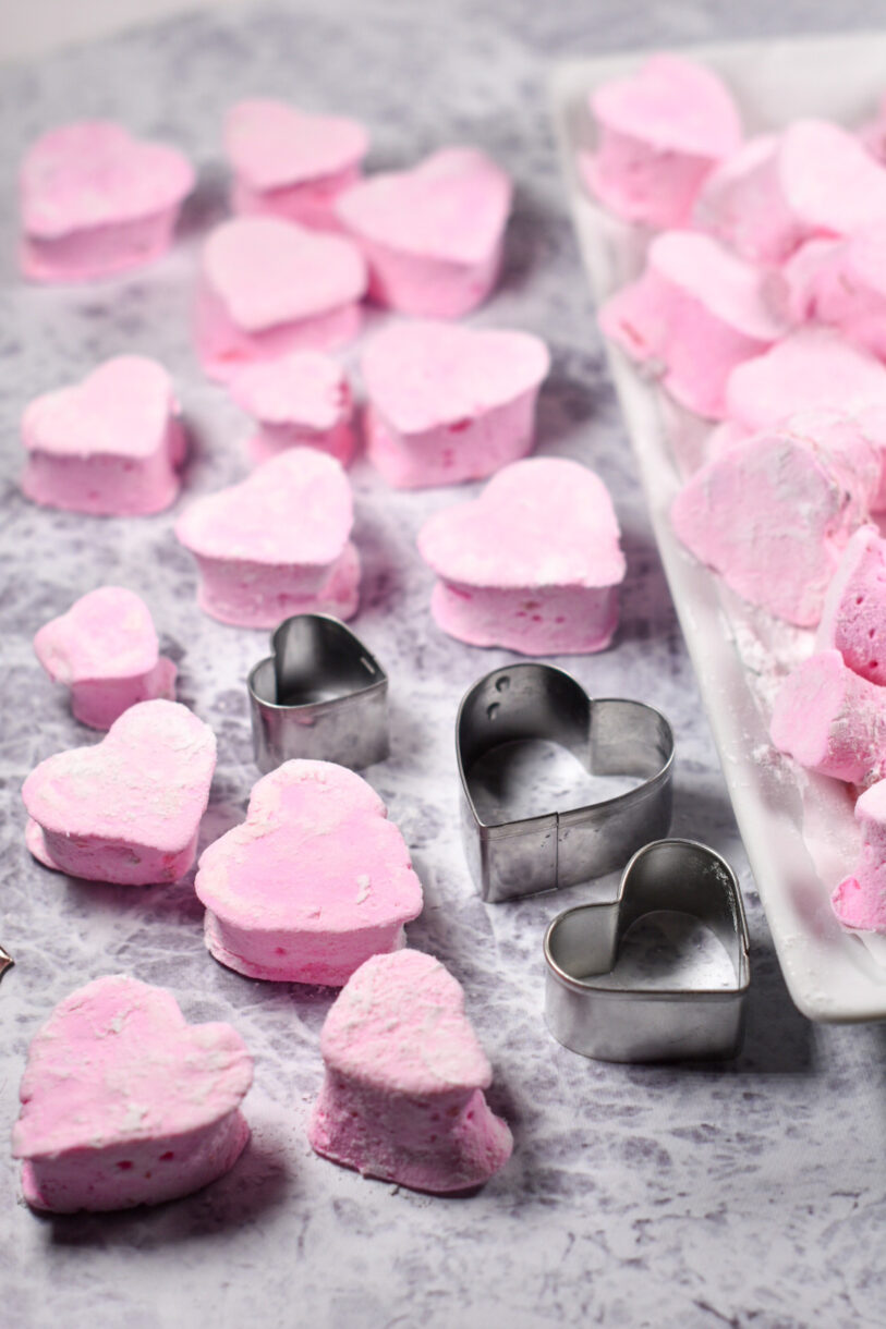 Homemade strawberry marshmallows and three metal heart cookie cutters ona grey textured surface