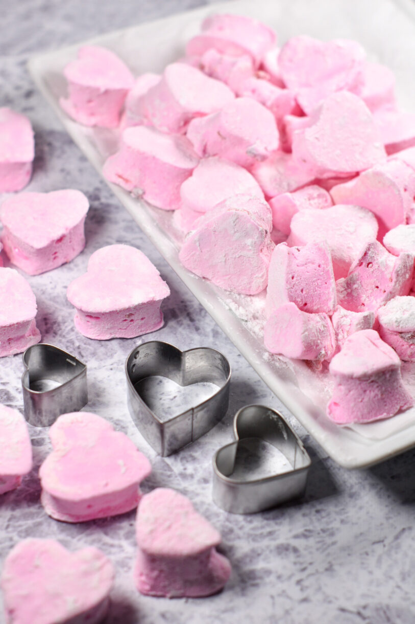 Homemade marshmallows shaped like hearts, arranged on a white rectangular tray alongside three heart shaped cookie cutters