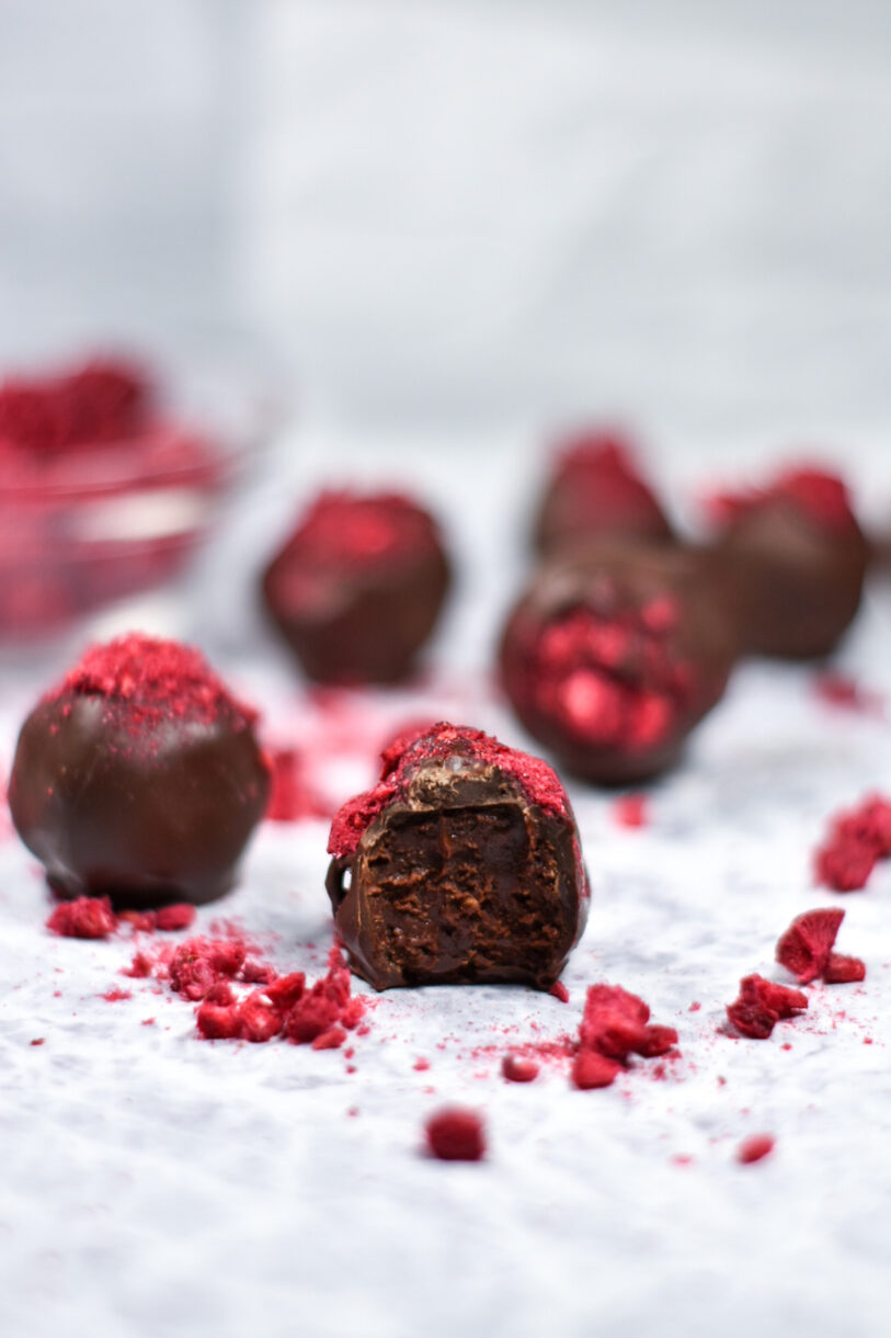 Chocolate raspberry truffle with a bite missing, surrounded by truffles and freeze-dried raspberries, on a white surface