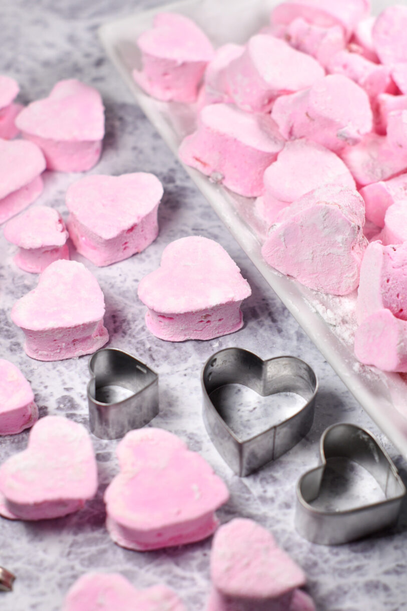 Homemade strawberry marshmallows and three heart shaped cookie cutters, with a tray of marshmallows in the background