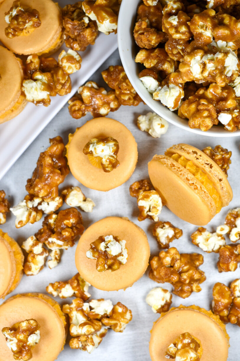 Looking down at a bowl of caramel corn, caramel corn macarons, and kernels of popcorn arranged on a white surface