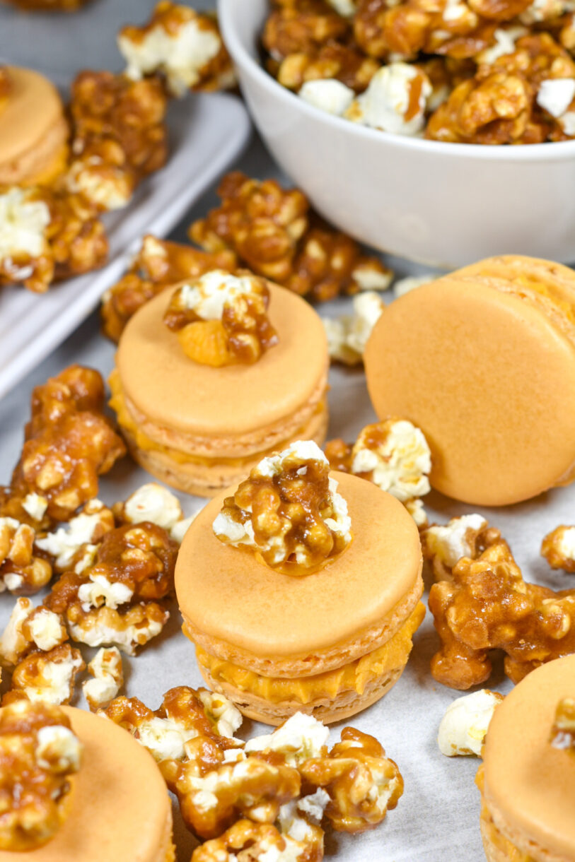 Caramel popcorn buttercream macarons surrounded by kernels of caramel corn, on a white surface