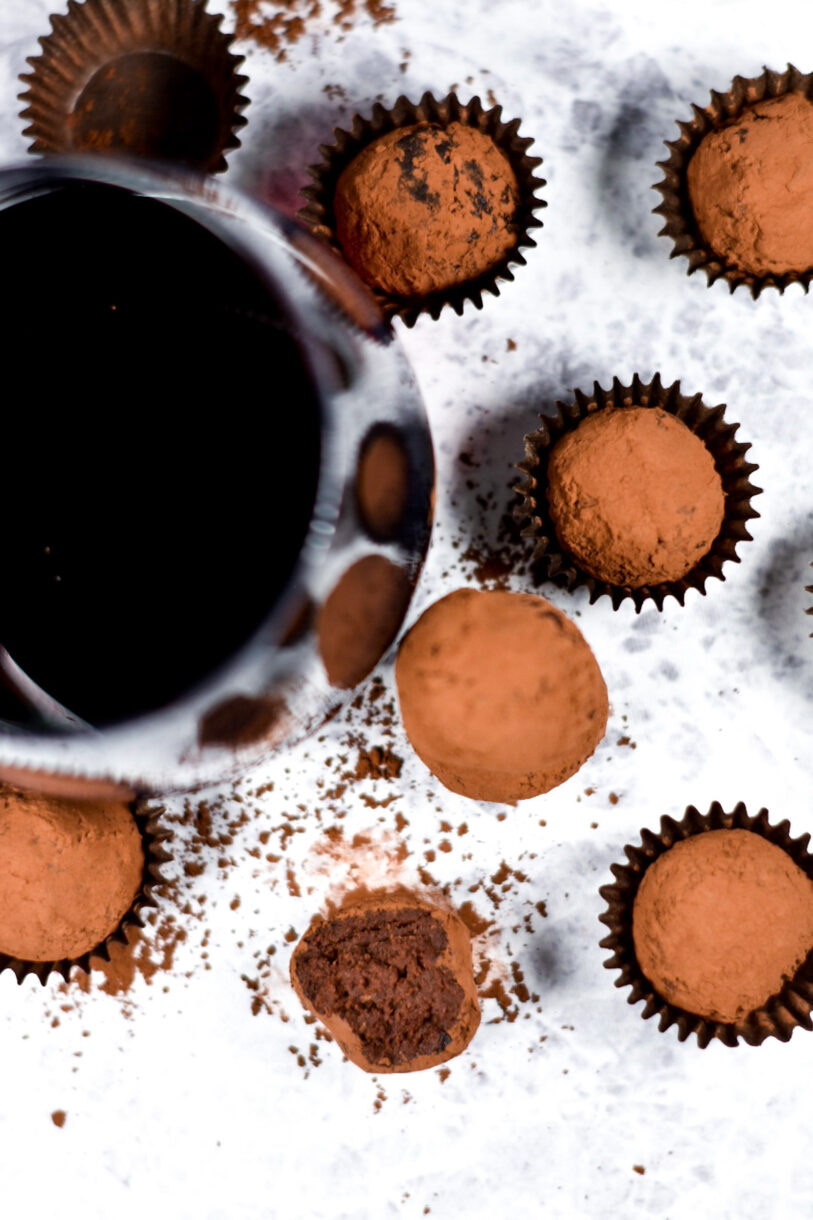 Red wine truffles, cocoa powder, and truffle cups, along with a stemless glass of red wine, on a white background
