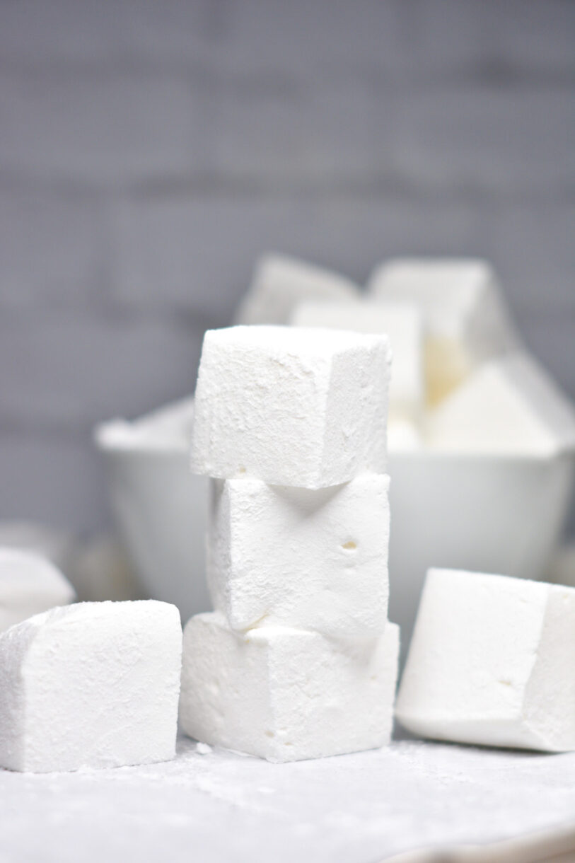 A stack of vanilla bean marshmallows with a white bowl of marshmallows in the background