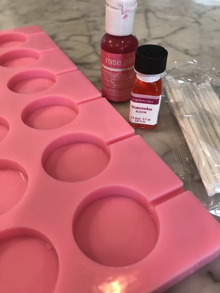 A silicone lollipop mold, a jar of watermelon flavoring, and rose pink food coloring