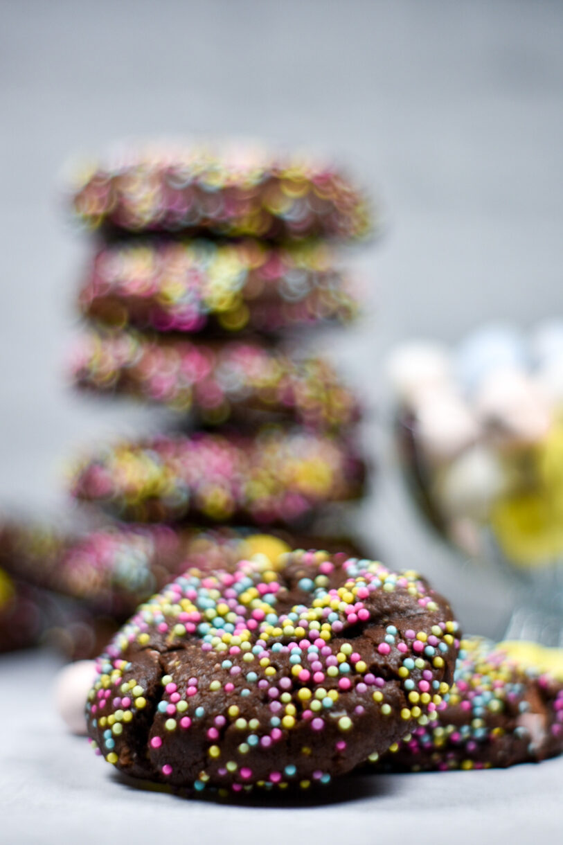 A nonpareil covered chocolate cookie in focus, with a stack of cookies blurry behind it