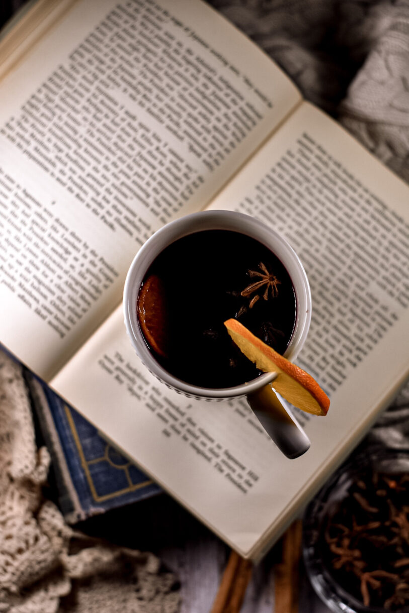 A mug of mulled red wine surrounded by books, cinnamon, and cozy fabrics