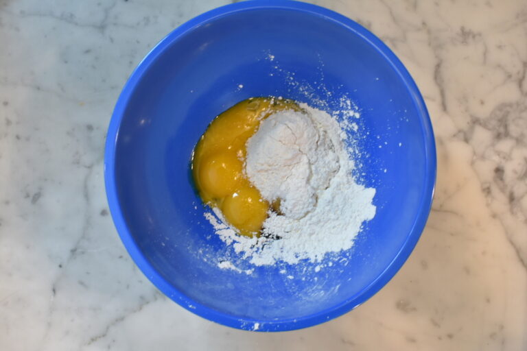 A blue mixing bowl with eggs, flour, and cornflour, sitting on a marble countertop