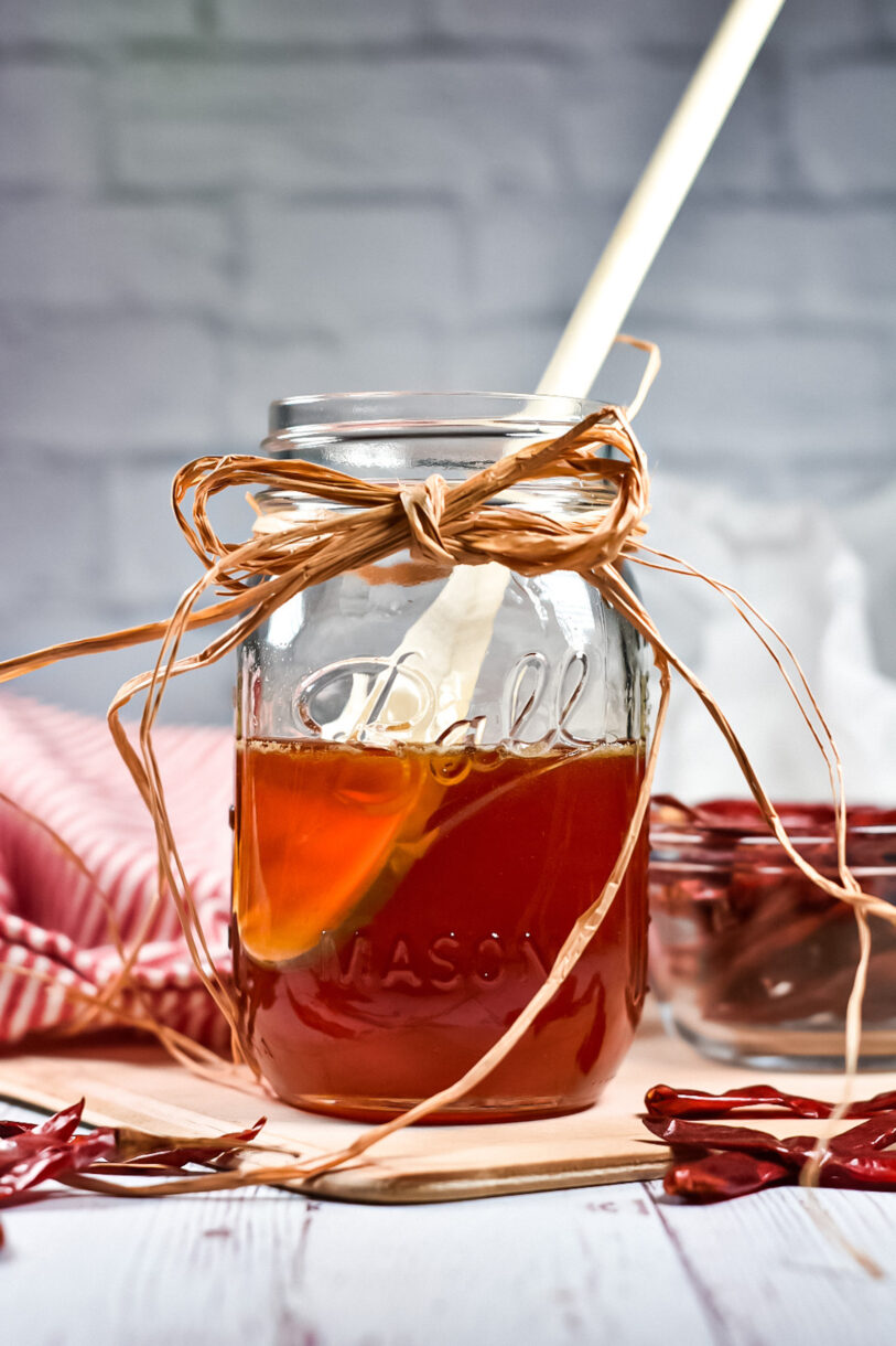 A glass jar of hot honey, along with dried chili peppers, wooden spoon, and red tea towel, on a white brick background