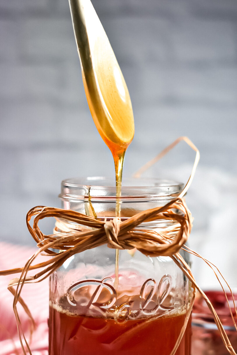 A wooden spoon drizzling hot honey into a Ball jar decorated with a twine bow