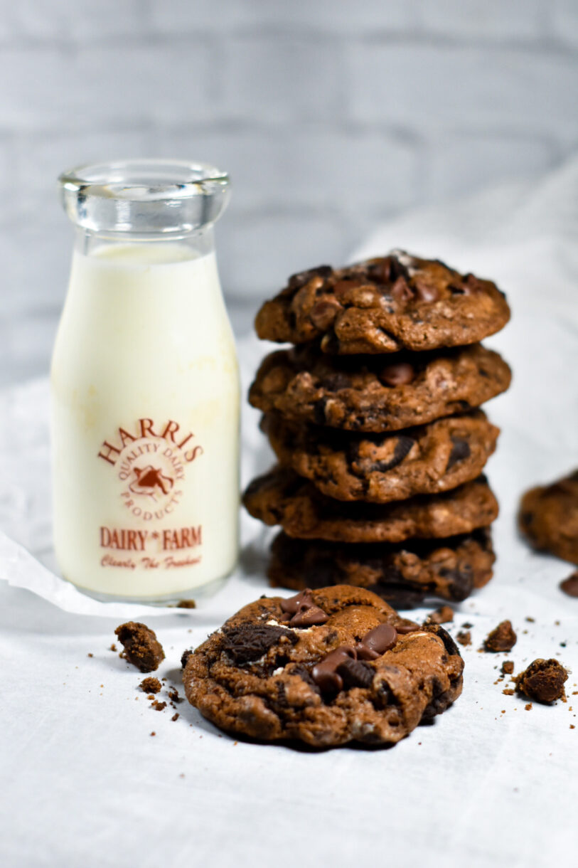 A stack of bakery-style chocolate oreo cookies and a small jug of milk sitting on a sheet of white parchment, in front of a white brick background