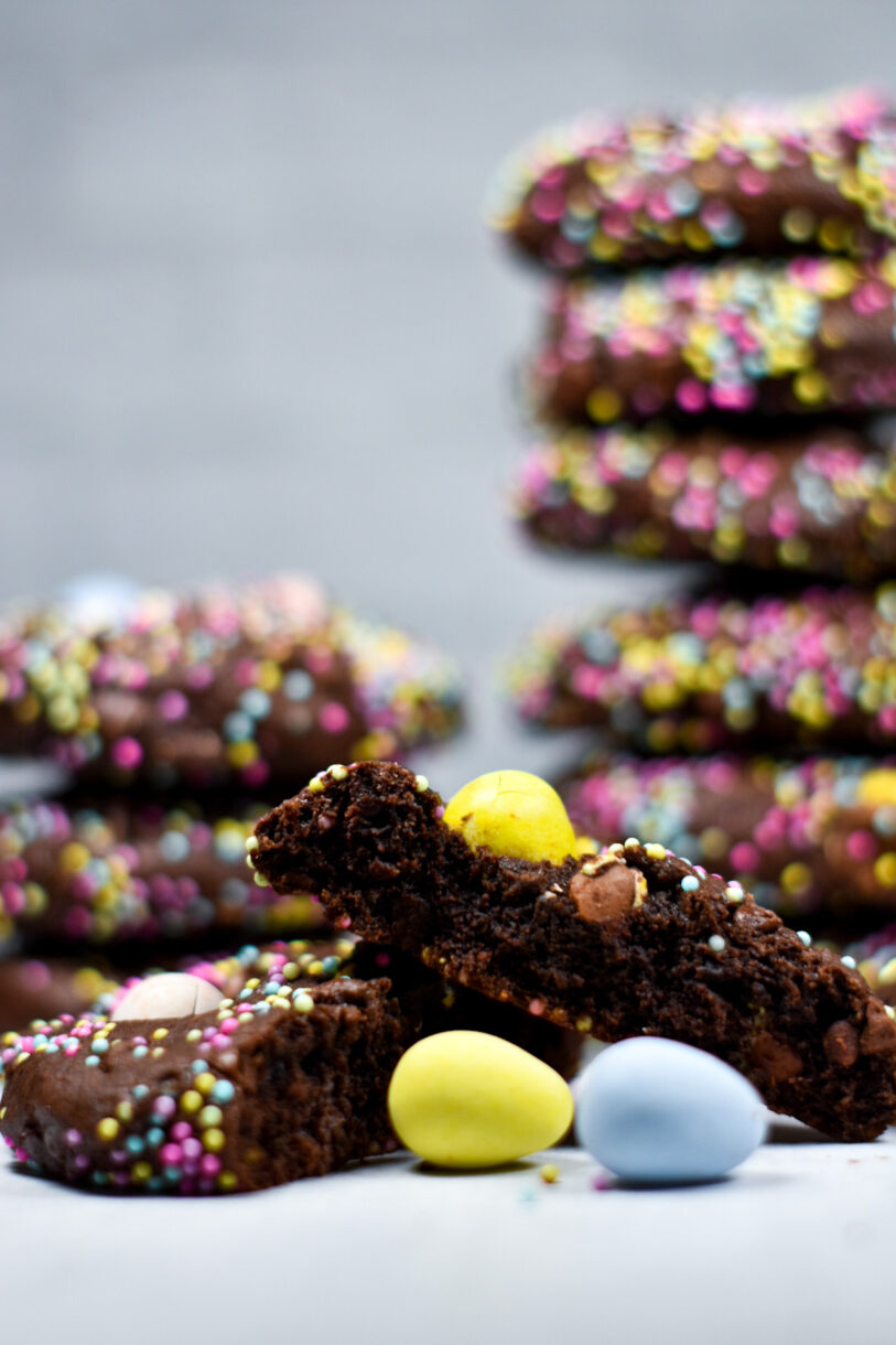 A sliced chocolate mini egg cookie, with a yellow and blue mini egg and a stack of cookies in the background