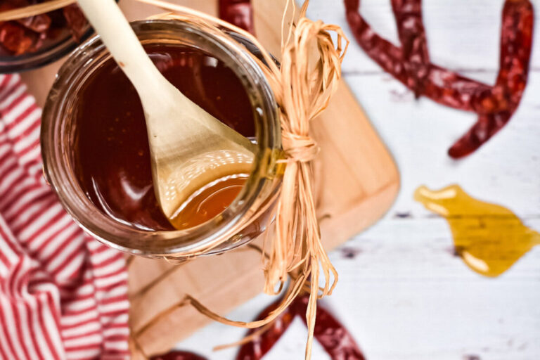 Looking down into a jar of hot honey which is sitting on a cutting board, with honey drizzled on the table and red chili pepper surrounding it