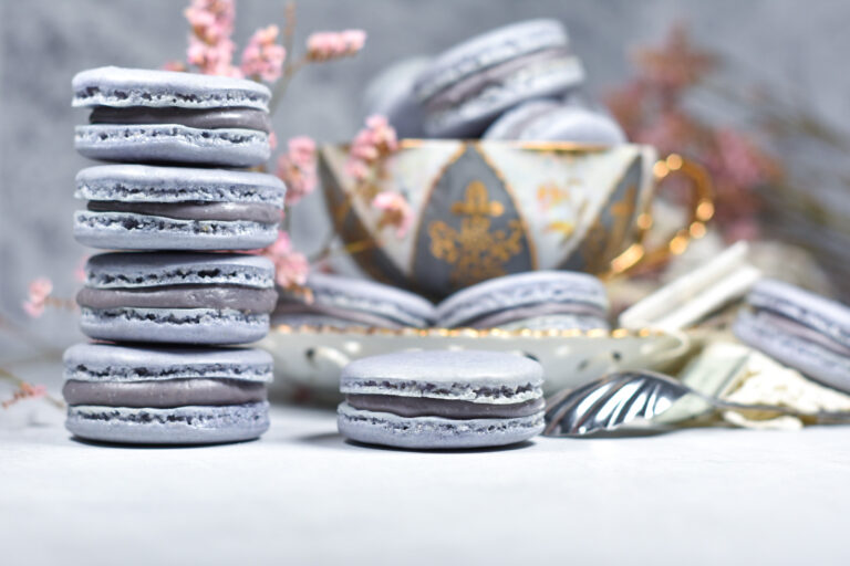 A stack of macarons next to a teacup filled with macarons, on a white background