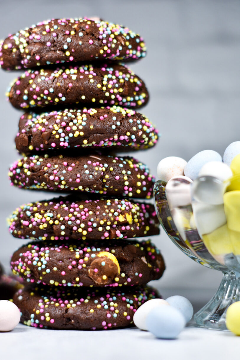 A tall stack of chocolate Easter cookies next to a glass bowl of mini eggs