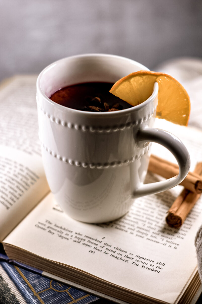A mug of red wine garnished with an orange wedge, sitting on top of an antique book with cinnamon sticks nearby