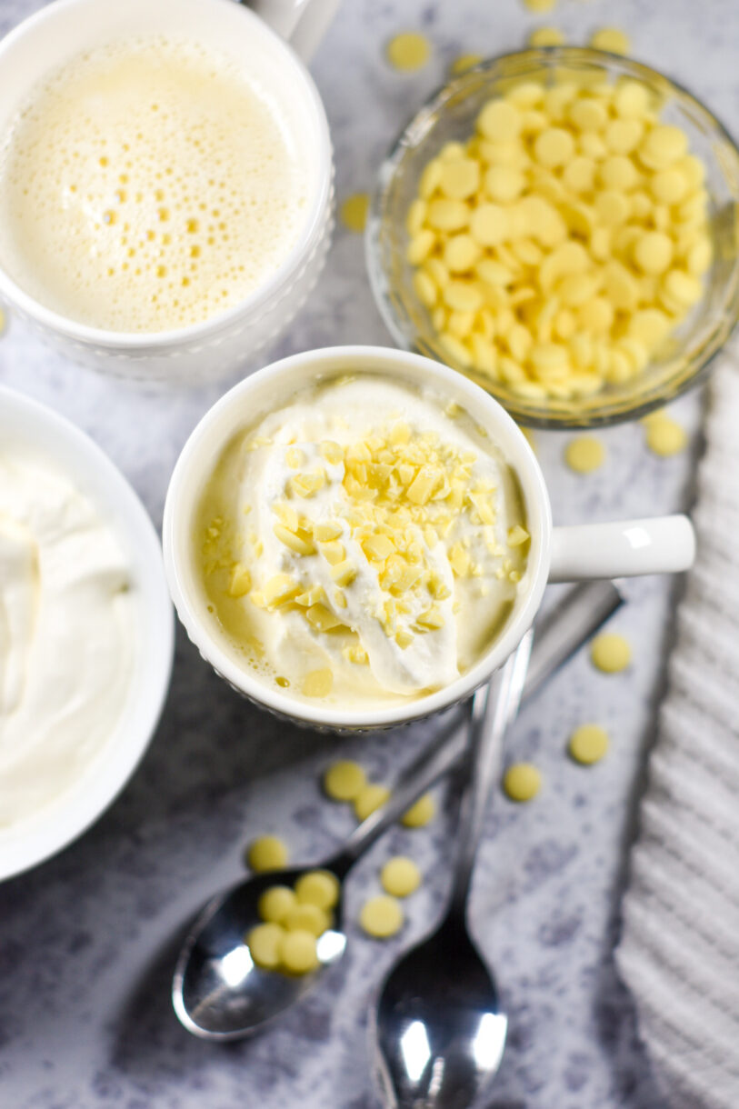 Mugs of white hot chocolate, two spoons, a bowl of whipped cream, and a bowl of white chocolate chips on a white background