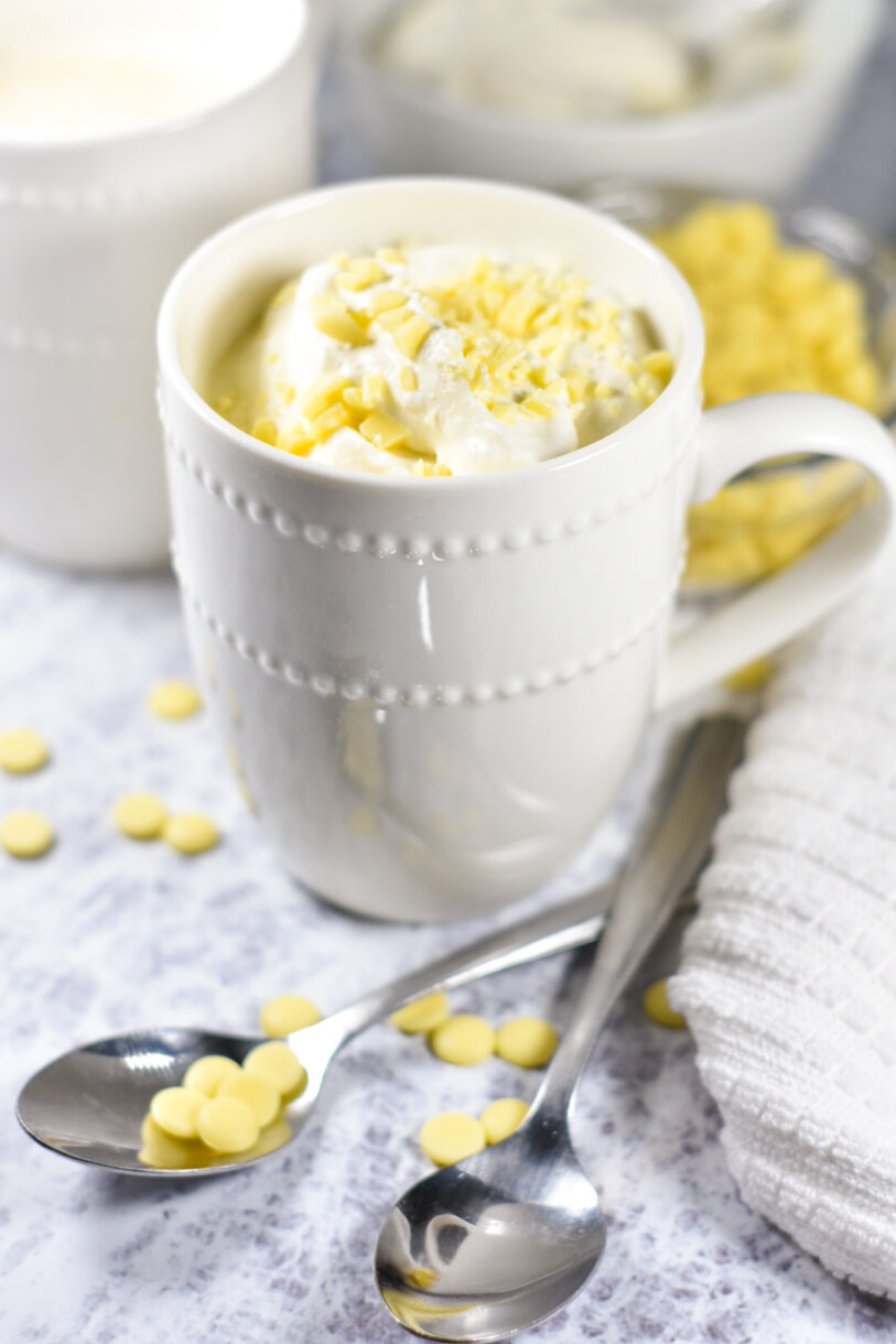 A white mug filled with white hot chocolate, two metal spoons, and white chocolate chips scattered on a white surface